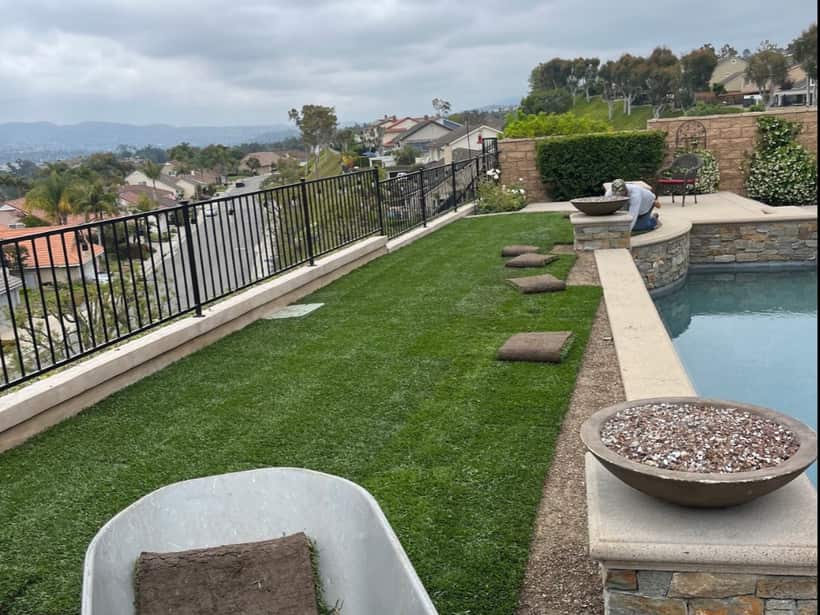 Artificial turf going in beside a hillside pool, rolls and weed barrier still on the ground